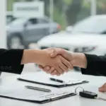 Pessoas apertando as mãos em uma mesa com documentos de financiamento de veículos e uma chave de carro, simbolizando um acordo bancário bem-sucedido.